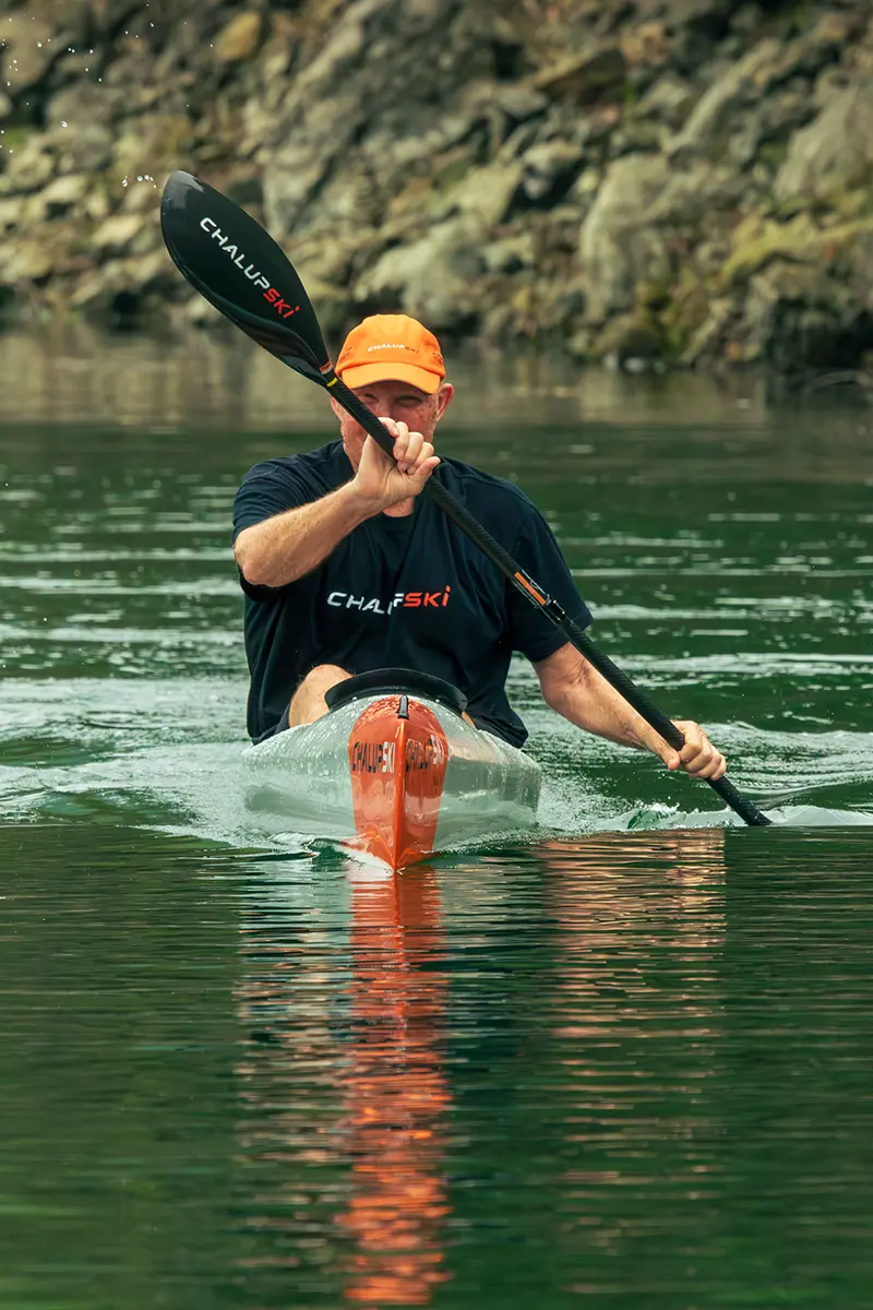 Un pagayeur en tenue ChalupSki sur une eau plate, le long d'une berge rocheuse.